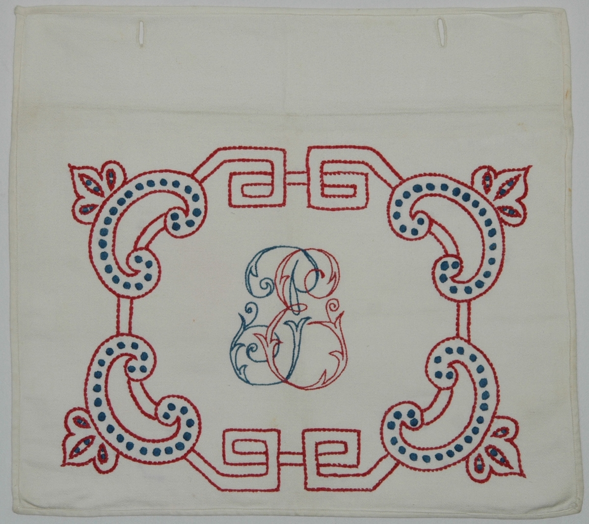 Av vit bomullsväv, s.k. angolaväv. Kuvertmodell med med övervikt lock åt baksidan knäppt med 2 linneknappar och knapphål, på framsidan initialerna PE omgivna av bård i "fornnordisk" stil. Sydd i stjälksöm och plattsöm med rött och blått bomullsgarn.