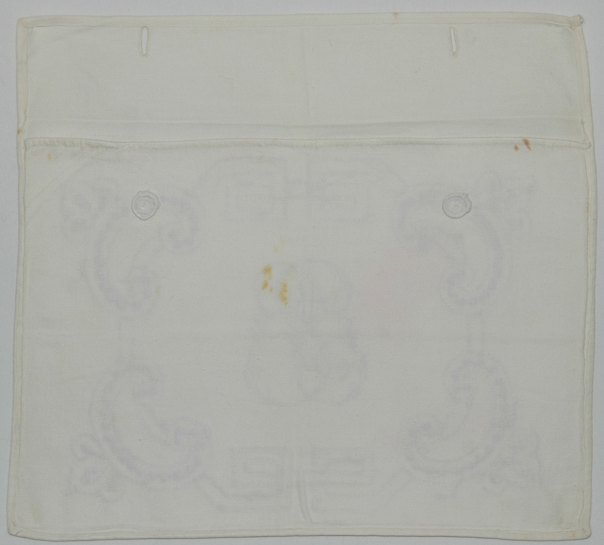 Av vit bomullsväv, s.k. angolaväv. Kuvertmodell med med övervikt lock åt baksidan knäppt med 2 linneknappar och knapphål, på framsidan initialerna PE omgivna av bård i "fornnordisk" stil. Sydd i stjälksöm och plattsöm med rött och blått bomullsgarn.