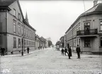 Klostergatan mot söder från Västra Storgatan i Jönköping. I gatukorsningen håller en poliskonstapel ett vakande öga på omgivningen. Längre in på gatan står en hästskjuts för utkörning av specerier, på vagnens baksida sitter reklam för bästa kaffet hos J. Lindstedt. Till vänster ligger Santessonska flickskolan och i bakgrunden syns Sofiakyrkans tornspira.