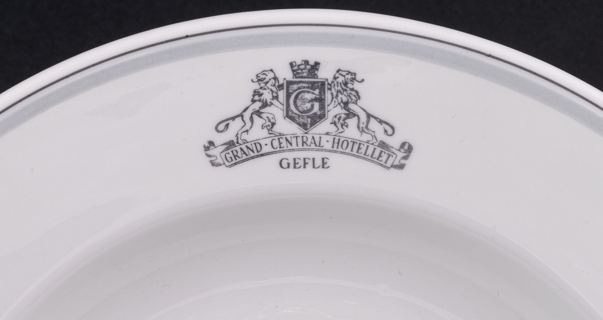 Tallrik, djup. Vit glasyr med ljusgrå och mörkgrå dekor. På brättet emblemet Grand Central Hotellet Gefle, samt två lejon hållande Gävle vapen. Ytterst på brättet två tunna grå ränder.