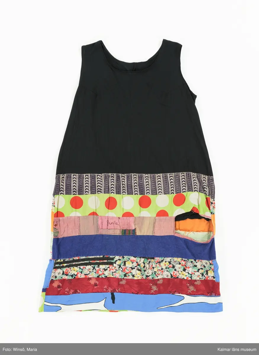 KLM 46157:589. Klänning. Ärmlöst konfektionsytt liv av svart trikå av konstmaterial. Kjol sydd av smala vågräta våder av nio olika mönstrade tyger, de flesta med tryckt mönster. Tygen är i huvudsak av bomull, något av konstmaterial. Ett rosa tyg är ihopskarvat av många små tygbitar, på en av bitarna står Gunila broderat i lila kedjestygn. Det ljusgröna tyget med röda och vita prickar finns även i kjolen till klänning KLM 46157:590. Ett tyg, svart botten med blommor i vitt, rött, gult och grönt, är klippt ur ett tidigare förkläde eller ev. flickklänning, fickan finns kvar.