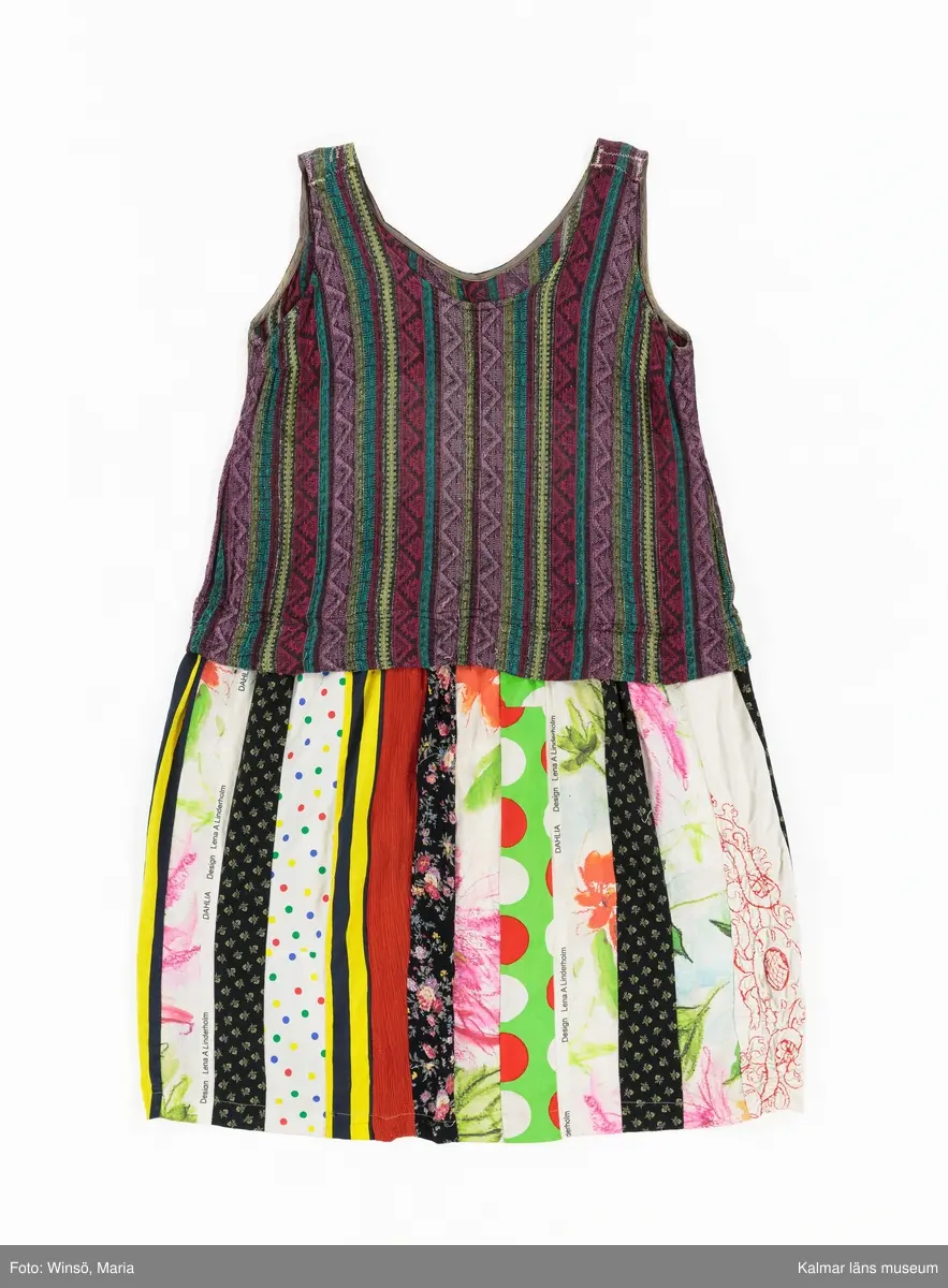 KLM 46157:590. Klänning. Ärmlöst liv av mönstervävt, trianglar och rutor, randigt bomullstyg i rött, rosa, grönt och gult mot svart botten. Halsringning och ärmöppningar kantade på insidan av beige snedslå. Kjol sydd av smala lodräta våder av åtta olika mönstrade tyger, de flesta med tryckt mönster. Tygen är i huvudsak av bomull. Ett tyg har broderat mönster i rött. Ett av de tryckta tygen är formgivet av Lena A Linderholm, mönstret heter Dahlia. Det svarta tyget med små tryckta blommor i gult finns även i kjolen till klänning KLM 46157:588. Det ljusgröna tyget med röda och vita prickar finns i kjolen till klänning KLM 46157:589.