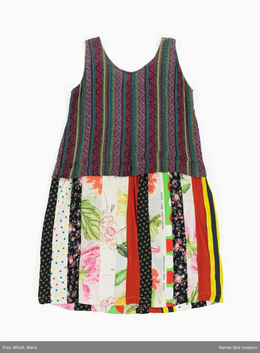 KLM 46157:590. Klänning. Ärmlöst liv av mönstervävt, trianglar och rutor, randigt bomullstyg i rött, rosa, grönt och gult mot svart botten. Halsringning och ärmöppningar kantade på insidan av beige snedslå. Kjol sydd av smala lodräta våder av åtta olika mönstrade tyger, de flesta med tryckt mönster. Tygen är i huvudsak av bomull. Ett tyg har broderat mönster i rött. Ett av de tryckta tygen är formgivet av Lena A Linderholm, mönstret heter Dahlia. Det svarta tyget med små tryckta blommor i gult finns även i kjolen till klänning KLM 46157:588. Det ljusgröna tyget med röda och vita prickar finns i kjolen till klänning KLM 46157:589.