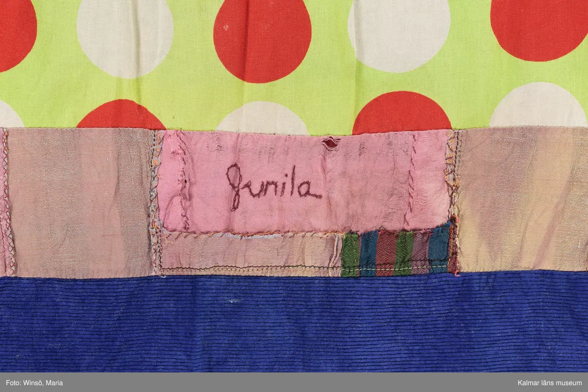 KLM 46157:589. Klänning. Ärmlöst konfektionsytt liv av svart trikå av konstmaterial. Kjol sydd av smala vågräta våder av nio olika mönstrade tyger, de flesta med tryckt mönster. Tygen är i huvudsak av bomull, något av konstmaterial. Ett rosa tyg är ihopskarvat av många små tygbitar, på en av bitarna står Gunila broderat i lila kedjestygn. Det ljusgröna tyget med röda och vita prickar finns även i kjolen till klänning KLM 46157:590. Ett tyg, svart botten med blommor i vitt, rött, gult och grönt, är klippt ur ett tidigare förkläde eller ev. flickklänning, fickan finns kvar.