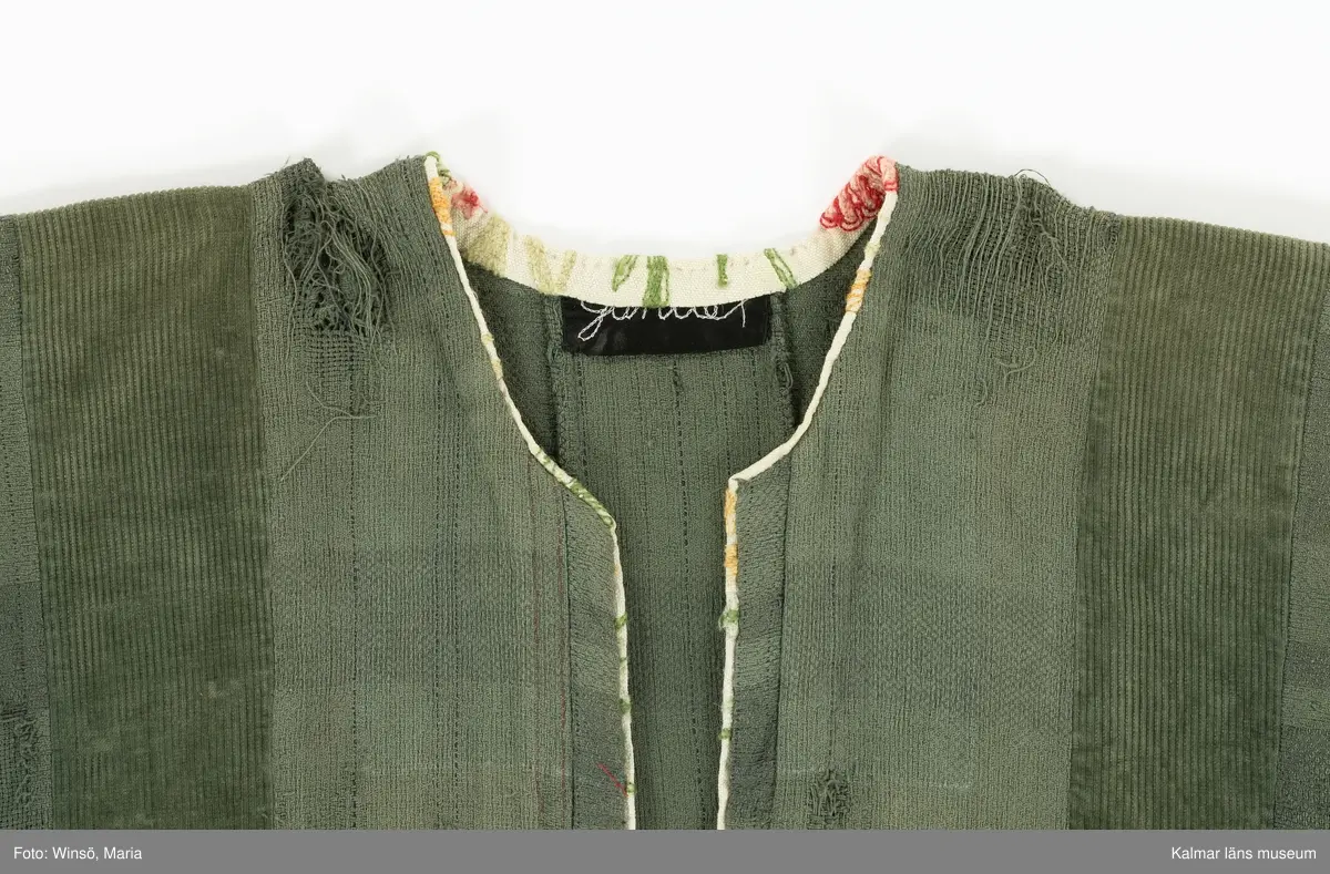 KLM 46157:591. Jacka. För dam. Grön. Rak modell med lång ärm. Sydd av två olika tyger, ett grövre bomullstyg med trådränder i blått och rött samt av bredspårig bomullsmanchester. Jackan är sydd av raka våder. Mellan några av våderna, vid nederkanten, isydda svarta kilar. Öppning mitt fram. Öppning och halsringning kantad med broderat, blommor i röda och gröna nyanser, ljust bommulstyg. Samma tyg är sytt i en bredare avslutning på ärmen. Rå kant vid jackans nederkant, sydd med glesa röda kaststygn. Handbroderad svart etikett på insidan i nacken med texten till stora delar dold av kantremsan. 
Tygerna i plagget är troligen återvunna. Det broderade tyget finns även i klänning KLM 46157:578.