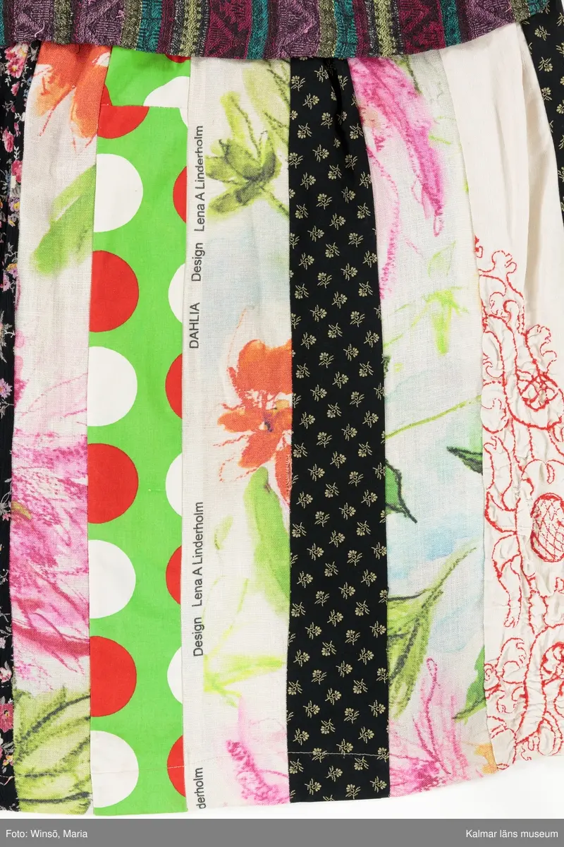 KLM 46157:590. Klänning. Ärmlöst liv av mönstervävt, trianglar och rutor, randigt bomullstyg i rött, rosa, grönt och gult mot svart botten. Halsringning och ärmöppningar kantade på insidan av beige snedslå. Kjol sydd av smala lodräta våder av åtta olika mönstrade tyger, de flesta med tryckt mönster. Tygen är i huvudsak av bomull. Ett tyg har broderat mönster i rött. Ett av de tryckta tygen är formgivet av Lena A Linderholm, mönstret heter Dahlia. Det svarta tyget med små tryckta blommor i gult finns även i kjolen till klänning KLM 46157:588. Det ljusgröna tyget med röda och vita prickar finns i kjolen till klänning KLM 46157:589.