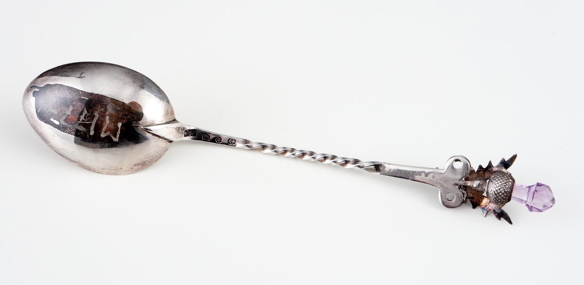 Kaffesked i silver.
Skeden har skaftet spiralvridet, avslutat med två ringar, två blad och en tistel. I skaftets yttre ände sitter en
fasettslipad, päronformad ljusviolett glassten. Skedbladet är förgyllt invändigt. 
Stämplar på skaftets baksida.

Inskrivet i huvudbok 1963