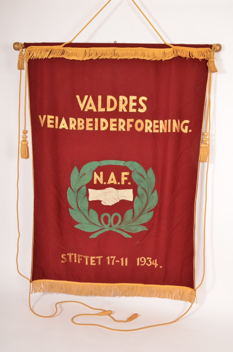 Fane i burgunder raudt ulltøy, lagt dobbelt. Teksten på fana er måla på med gullfarge. Dei andre motiva (bladkransen og hendene) er også måla på tøyet. Kanten i øvre- og nedrekant er påsett gullfarga frynse-bord. Langs sidene er det gullsnor med duskar. På baksida er teksten også måla på med gullfarge. 
Opphengt i øvre i kanten med ei gullmåla trestang med dreia kuler i kvar ende. Denne er tredd inn i ein løpgang. Krok i metall midt på stanga.