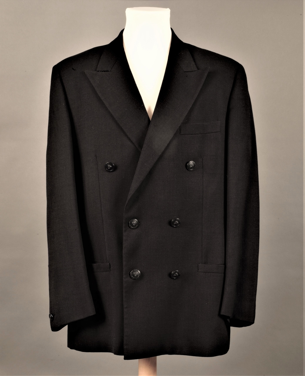 Hoftelang, dobbeltspent, med 6 metallknapper, med Versace-husets "Istante"-logo - Zevs hode - i relieff. Lange slag, og kun de nederste 4 knapper skal kneppes. Lapellene (slagene) er spisse og lengre enn kragen. Ett ekstra knapphull for innvendig knapping, men knappen på innsiden av venstre forstykke mangler. To-søms ermer med falsk splitt, lukket knapphull og pynteknapp. Horisontal lomme med belegg på hver hofte og stikklomme med belegg på venstre bryst. Innvendig, passepoilert lomme på hvert bryst, den høyre lukket med klaff og usynlig knapp. Søm midt bak og splitt nederst i hver panelsøm bak. Forstykkene er skåret rettvinklet i nederkant midt foran. Helfôret med svart fôrsilke. Kragestanden og kragen er kledd med filtaktig materiale på baksiden og jakken har puter i skuldrene.