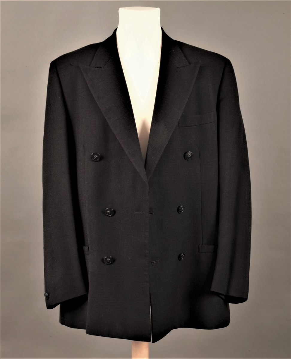 Hoftelang, dobbeltspent, med 6 metallknapper, med Versace-husets "Istante"-logo - Zevs hode - i relieff. Lange slag, og kun de nederste 4 knapper skal kneppes. Lapellene (slagene) er spisse og lengre enn kragen. Ett ekstra knapphull for innvendig knapping, men knappen på innsiden av venstre forstykke mangler. To-søms ermer med falsk splitt, lukket knapphull og pynteknapp. Horisontal lomme med belegg på hver hofte og stikklomme med belegg på venstre bryst. Innvendig, passepoilert lomme på hvert bryst, den høyre lukket med klaff og usynlig knapp. Søm midt bak og splitt nederst i hver panelsøm bak. Forstykkene er skåret rettvinklet i nederkant midt foran. Helfôret med svart fôrsilke. Kragestanden og kragen er kledd med filtaktig materiale på baksiden og jakken har puter i skuldrene.