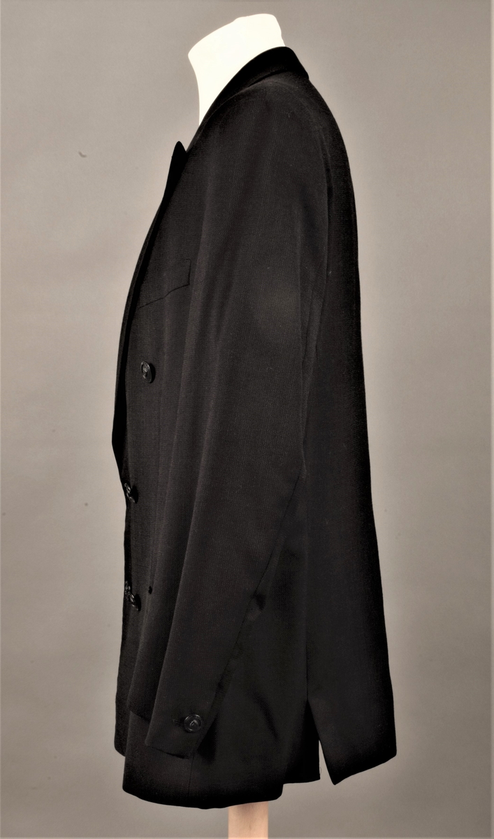 Hoftelang, dobbeltspent, med 6 metallknapper, med Versace-husets "Istante"-logo - Zevs hode - i relieff. Lange slag, og kun de nederste 4 knapper skal kneppes. Lapellene (slagene) er spisse og lengre enn kragen. Ett ekstra knapphull for innvendig knapping, men knappen på innsiden av venstre forstykke mangler. To-søms ermer med falsk splitt, lukket knapphull og pynteknapp. Horisontal lomme med belegg på hver hofte og stikklomme med belegg på venstre bryst. Innvendig, passepoilert lomme på hvert bryst, den høyre lukket med klaff og usynlig knapp. Søm midt bak og splitt nederst i hver panelsøm bak. Forstykkene er skåret rettvinklet i nederkant midt foran. Helfôret med svart fôrsilke. Kragestanden og kragen er kledd med filtaktig materiale på baksiden og jakken har puter i skuldrene.