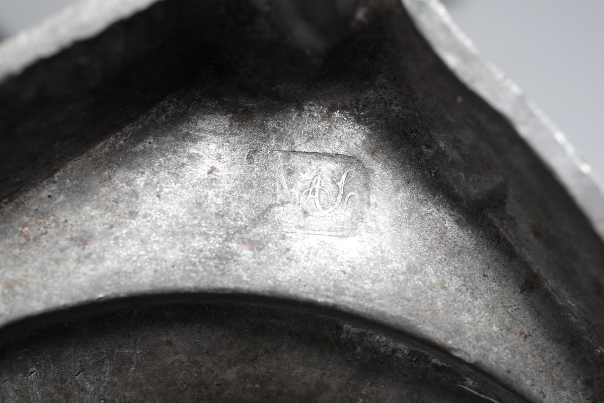 Ljusstake av tenn. 
S.k. kolonnmodell med fyrkantig kannelerad och pärlstavsdekorerad bottenplatta, på fyra linsfötter. 
Stämplar (mycket nötta och svårtydda) på fotplattans undersida.

Inskrivet i huvudbok 1955.