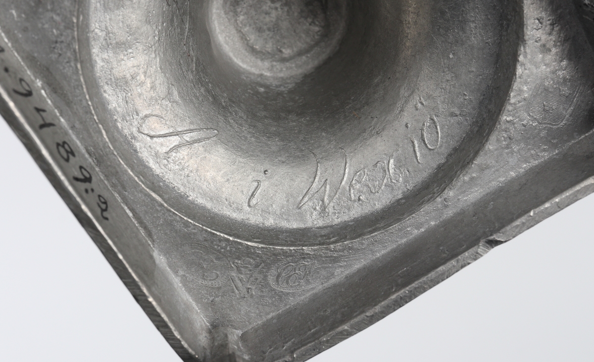Ljusstake av tenn.
Empire. Räfflad urnpipa på klockat postament. Fyrkantig fotplatta med blombård, försedd med fyra kulfötter. 
Stämplar i botten + gjuten signering: "A i Wexiö".

Inskrivet i huvudbok 1937.