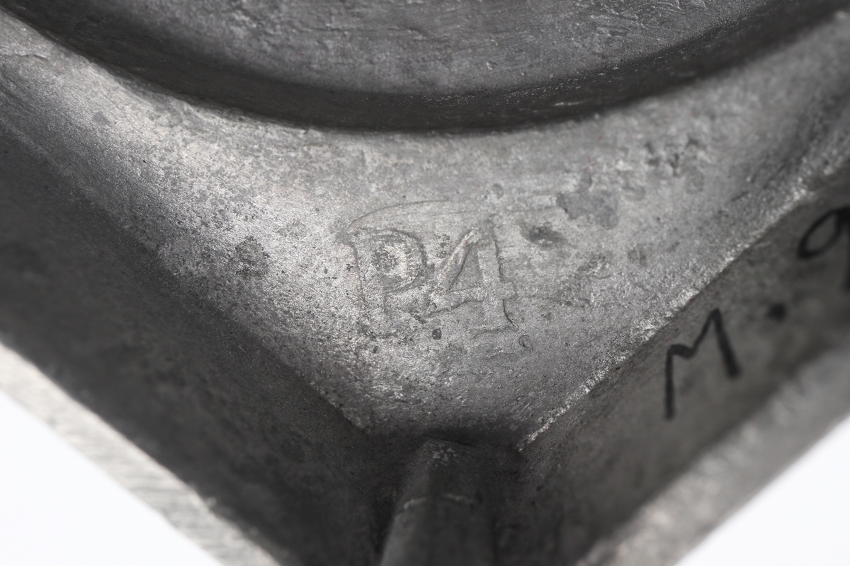 Ljusstake av tenn.
Empire. Räfflad urnpipa på klockat postament. Fyrkantig fotplatta med blombård, försedd med fyra kulfötter. 
Stämplar i botten + gjuten signering: "A i Wexiö".

Inskrivet i huvudbok 1937.