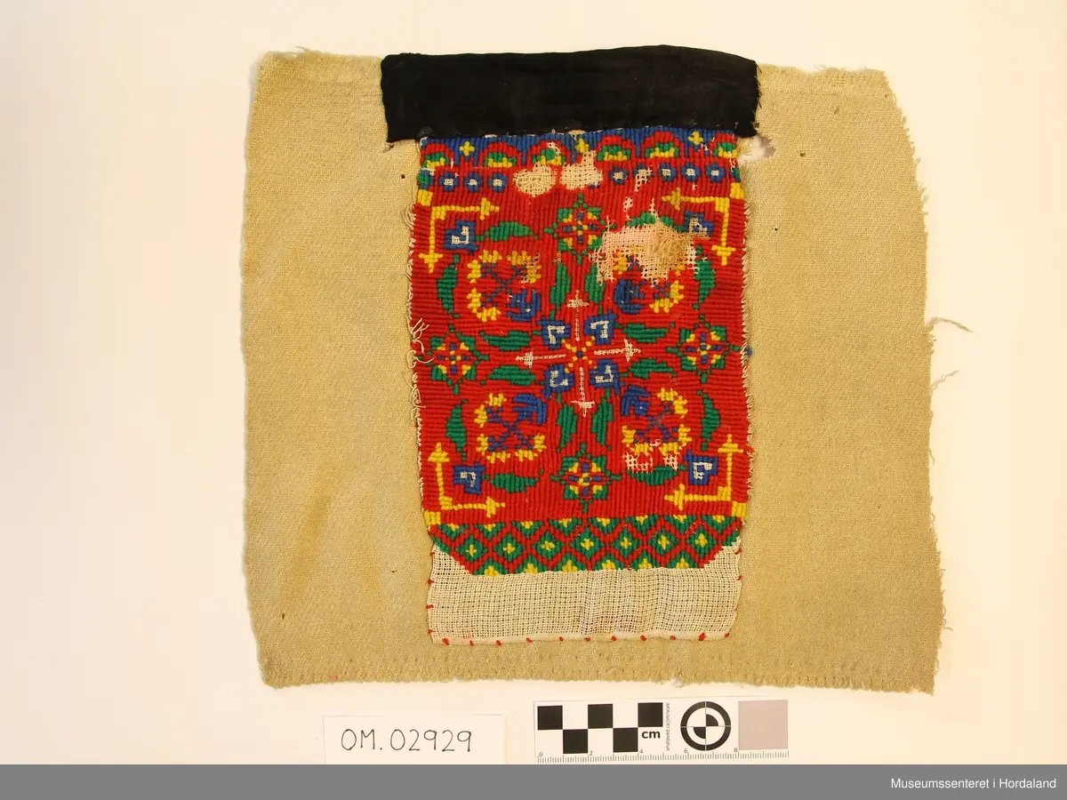 Rektangulært broderi på kvadratisk bakstykke. Plattsaum (halve kross-sting) i fleire retningar sydd på stramei. Nokre usydde og utslitte område. Raud hovudfarge. Diverse geometriske figurar og ei slags åttebladrose i midten. Bord nede  og oppe. Broderistoffet går litt nedom den nederste borda. Svart kant oppe. Alt er sydd på eit ullstoff vove i kypert. Fleire hol.