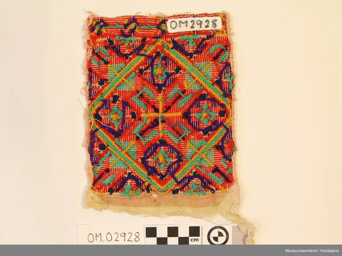 Rektangulær bringeduk med "enkel" og dobbel kross-sting i forskjellige retningar. Stor åttebladrose i midten og fire små oppe, nede og på sidene med ei rame rundt. I kvart hjørne er det eit blad av åttebladrose. Bord oppe med anna mønster (slags stjernemønster).  Raud hovudfarge. Dei mørkeblå krossane er sydd med kross-sting. Fargane er litt falma, spesielt den grønturkise og gule.