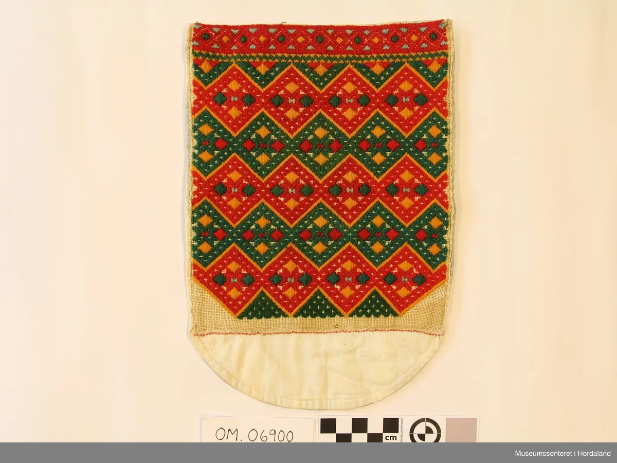 Brodert brystduk i repeterande mønster med sikksakk-border. Broderiet er rektangulært, bakstykket endar i ein halvrunding nedom broderiet. Stripete stoff på baksida.