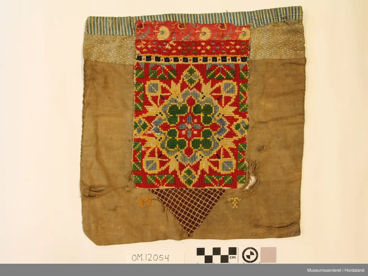 Hovudmotivet på bringeduken er brodert i krossting på stramei med ullgarn, det kvite er brodert med bomullsgarn.  Oppe er det ei smal borde i smøyg.Ovanfor der att er det ei borde klipt av eit raudt trykt stoff (Kattuntrykk). Dette er montert på eit bakstykke av bomullslerret som kanskje har vore kvitt, er brunt no.. Under hovudmotivet er det ein liten brunrutete trekant i eit anna bomullstoff. På kvar side av denne trekanten står initialane, M til venstre og T til høgre. Øvst oppe er det ein kant av eit blå-  og kvitstripete bomullststoff som er bretta rundt og går nedatt på baksida. Rett nedanfor det er det ein anna stoff.