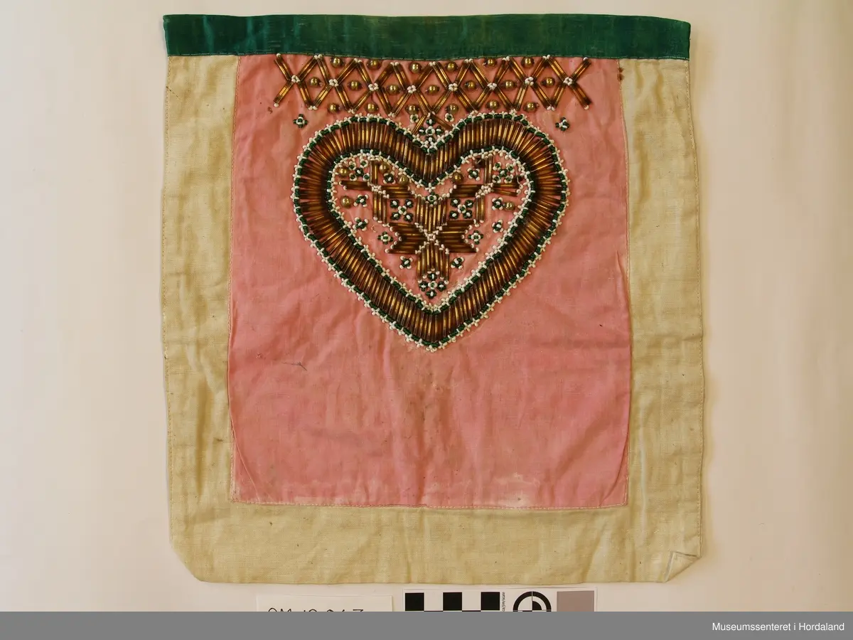 Perlebringeduk, der hovudmotivet er eit hjarta med ei 8-bladrose inni, alt laga av forgylte stråperler. Rundt kanten på hjarta er det border med kvite og grøne småperler. Ovanfor hjarta er der ei borde med stråperler og forgylte store perler innimellom. Alt er montert på eit rosa bomullslerret (har det vore raudt?) Oppe på kanten er det sydd eit grønt silkeband. Bakstykker dobbelt kvitt bomullslerret.