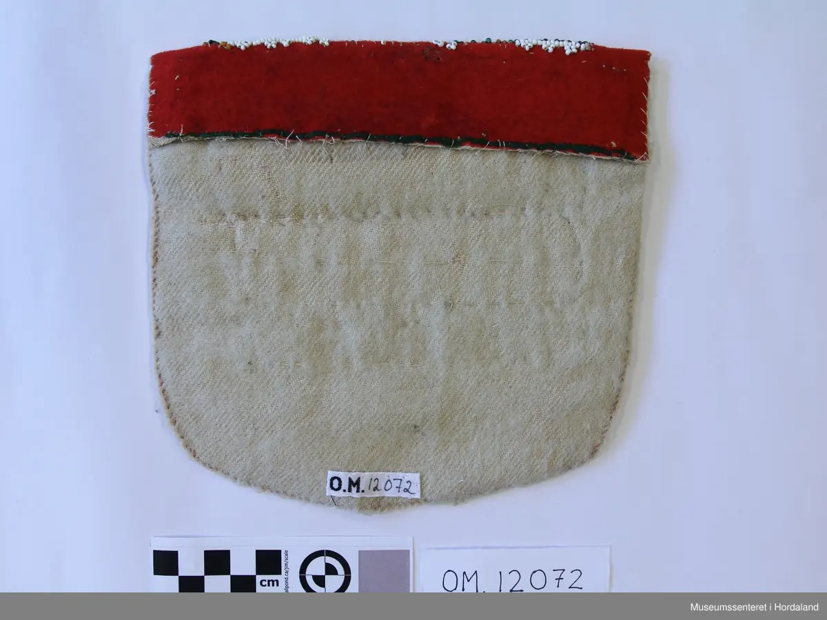 Bringeduk med mønster i border montert på raudt vadmål, annakvar perleborde og gullagraman 4 til saman, bakstykke av kvitt vadmål. Oppe er bringeduken bretta ned der som det har vore ein grøn kant; det viser restar av det grøne stoffet. Den øverste perlekanten er akkurat i bretten.