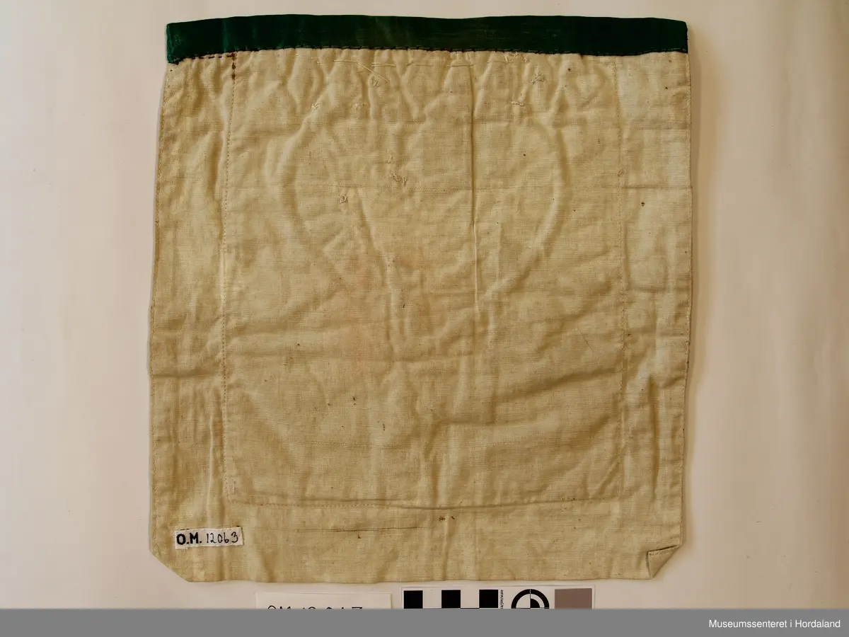 Perlebringeduk, der hovudmotivet er eit hjarta med ei 8-bladrose inni, alt laga av forgylte stråperler. Rundt kanten på hjarta er det border med kvite og grøne småperler. Ovanfor hjarta er der ei borde med stråperler og forgylte store perler innimellom. Alt er montert på eit rosa bomullslerret (har det vore raudt?) Oppe på kanten er det sydd eit grønt silkeband. Bakstykker dobbelt kvitt bomullslerret.