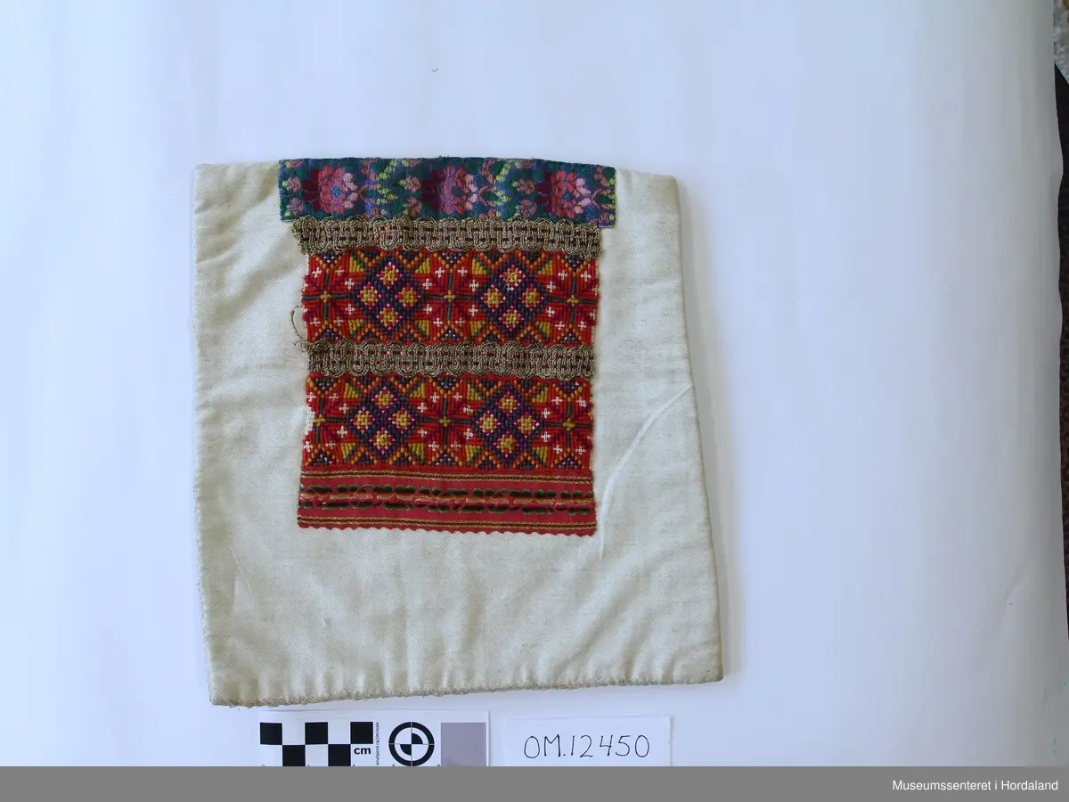 Bringeklut med ullbroderi i korssting i raud, grønt, gult og lilla. Sydd på stramei. Metallborder innimellom broderiet. Rosete silkevevnad oppe i blått,grønt og fleire raudlilla fargar. Eit silkeband nede i rosa, grøn og gul farge.  Montert på kvitt bomullsstoff.