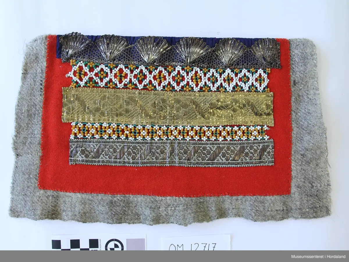 Rektangulær bringeduk med 2 rader perlenett montert på raudt stoff - annakvar perlerad og metallband. Både sølv og gull metallband. Kronekvarde oppe montert på blått band.  Bredda er større enn høgda. Bakstykke er eit vove ullstoff i kypert.