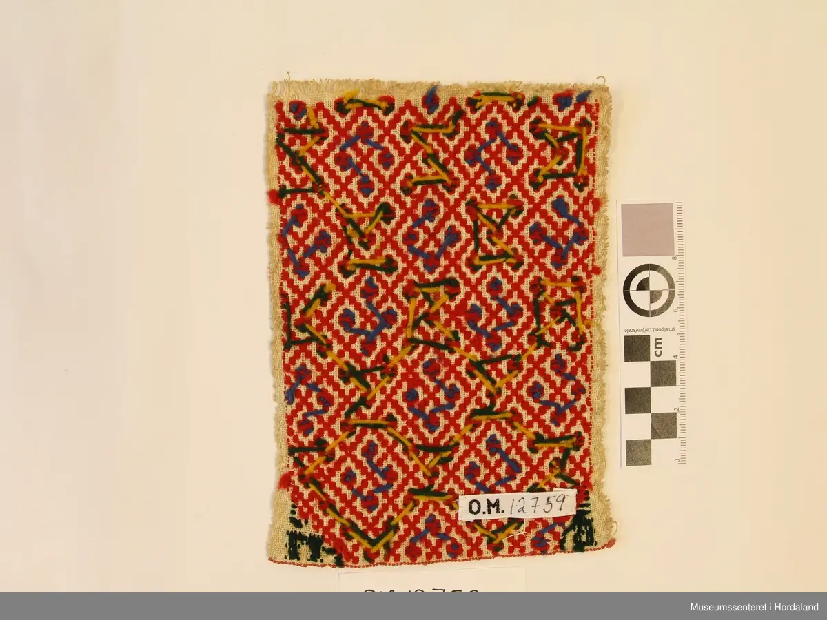 Broderi i plattsaum. Motivet er småruter. Nede i kvart hjørne er det brodert B i venstre hjørne og M i høgre hjørne. Brodert på kvitt linstoff.