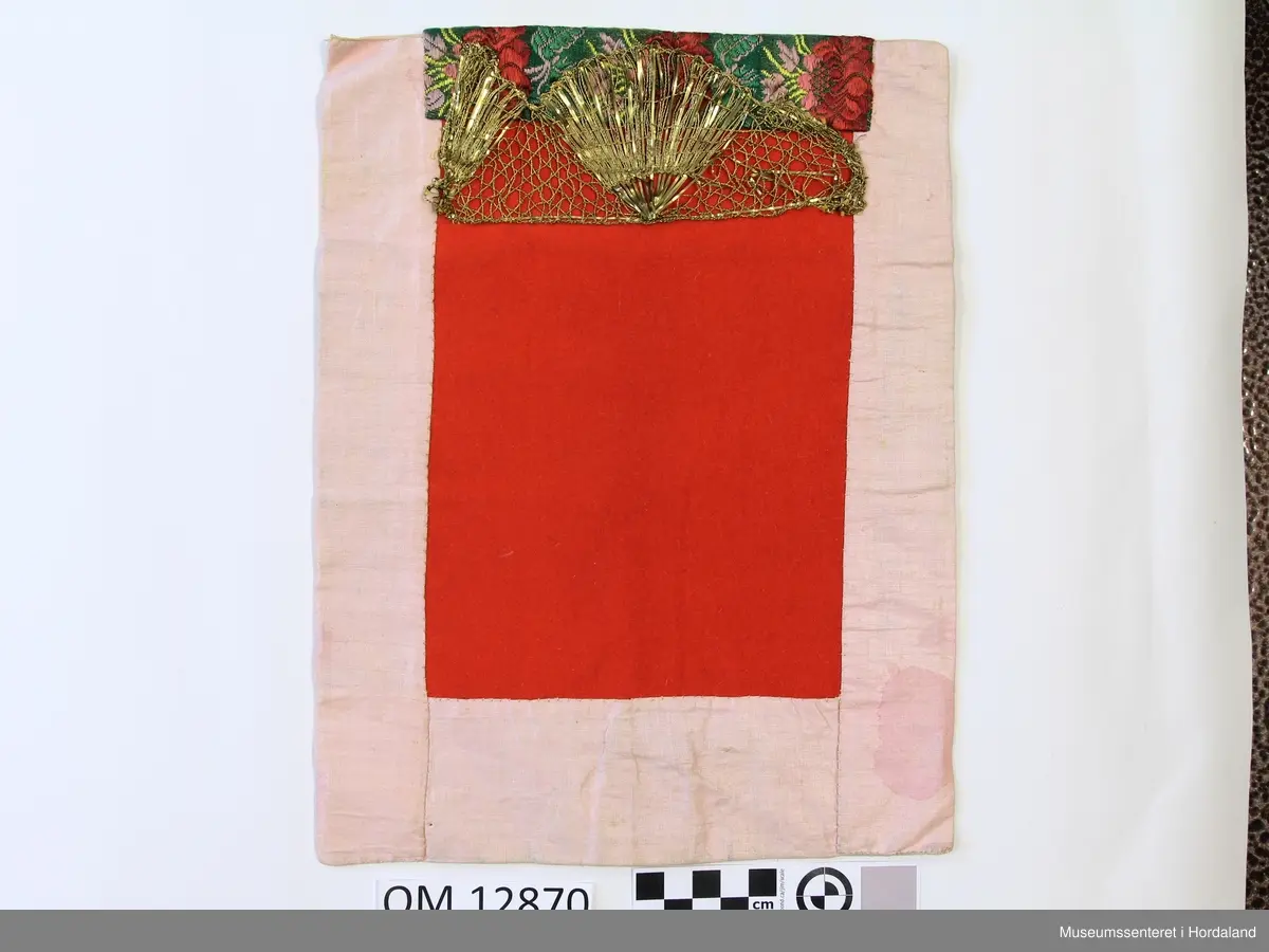 Rektangulær bringeduk med raudt ullstykke framme uten brodering. Oppe er det ein kant med gullfrga metallbord oppå eit brokadeband, metallborden går og eit stykke ned på det raude stoffet. Rosa, blankt fõrstoff til brei kanting og bakstykke. Alt er handsauma. Eit stykke papir el l inni til forsterking. Litt felkkete på forstoffet.