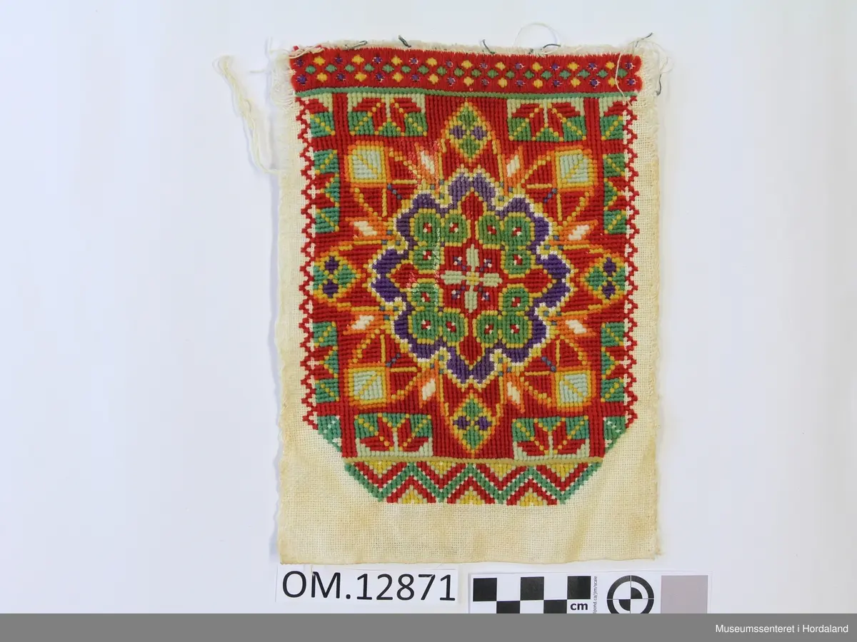 Rektangulær brystduk brodert på eit vevd på eit bomullstoff. Plattsaum (halve kross-sting). Blomstermotiv i midten sydd i fleire retningar. Brodert bord oppe og nede. Raud er hovudfargen.