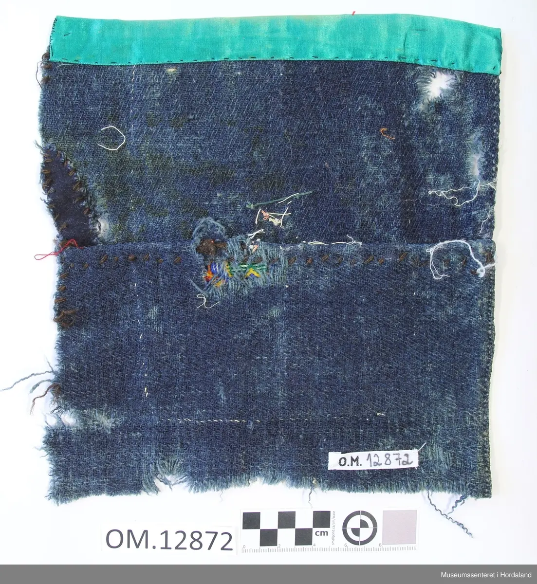 Rektangulær bringeduk på kvadratisk blått, kypertvevd bakstykke i ull. Broderiet er sydd på stramei. Blomstermotiv i midten og diverse geometriske figurer. Bord oppe og nede. Plattsaum og enkel kross-sting i fleire retningar. fire store glasperler inne i blomsten- Brodert i kross-sting under broderiet A M D T. Turkis band øvst oppe på kanten, eit raudt ullstoff imellom den turkise kanten og broderiet. Holete. Bakstykket er sydd saman av to deler og lappa. Kant lagt inn på eine sida.