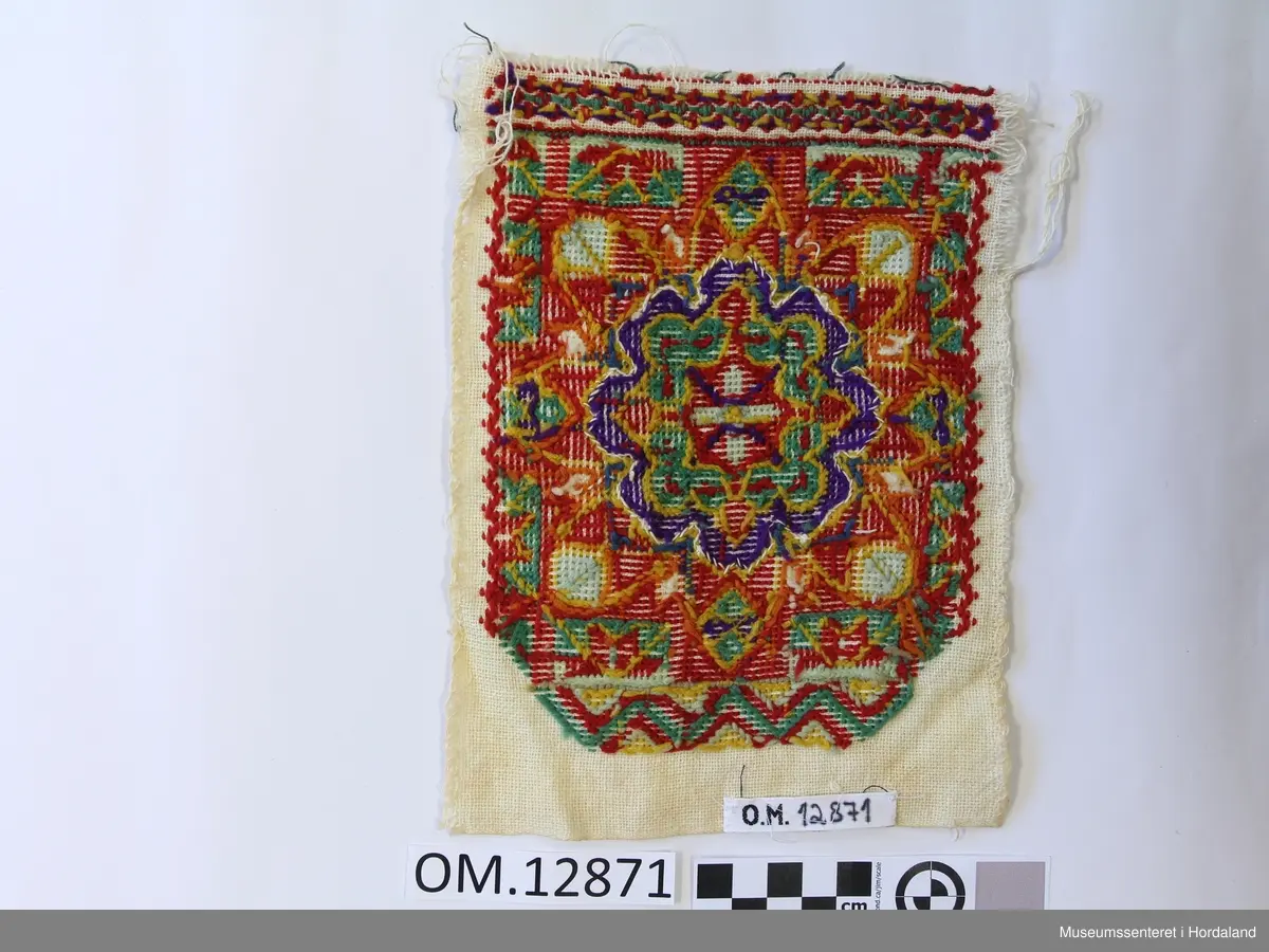 Rektangulær brystduk brodert på eit vevd på eit bomullstoff. Plattsaum (halve kross-sting). Blomstermotiv i midten sydd i fleire retningar. Brodert bord oppe og nede. Raud er hovudfargen.