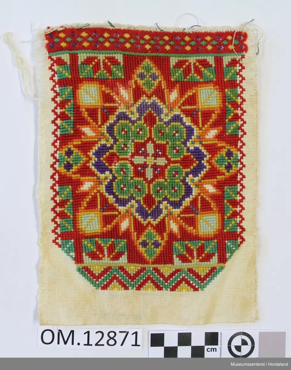 Rektangulær brystduk brodert på eit vevd på eit bomullstoff. Plattsaum (halve kross-sting). Blomstermotiv i midten sydd i fleire retningar. Brodert bord oppe og nede. Raud er hovudfargen.