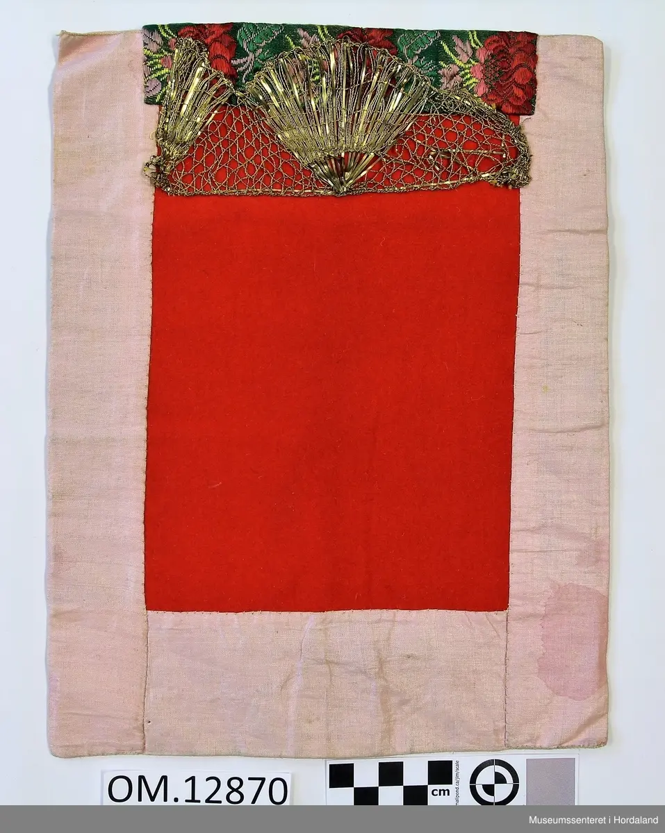 Rektangulær bringeduk med raudt ullstykke framme uten brodering. Oppe er det ein kant med gullfrga metallbord oppå eit brokadeband, metallborden går og eit stykke ned på det raude stoffet. Rosa, blankt fõrstoff til brei kanting og bakstykke. Alt er handsauma. Eit stykke papir el l inni til forsterking. Litt felkkete på forstoffet.