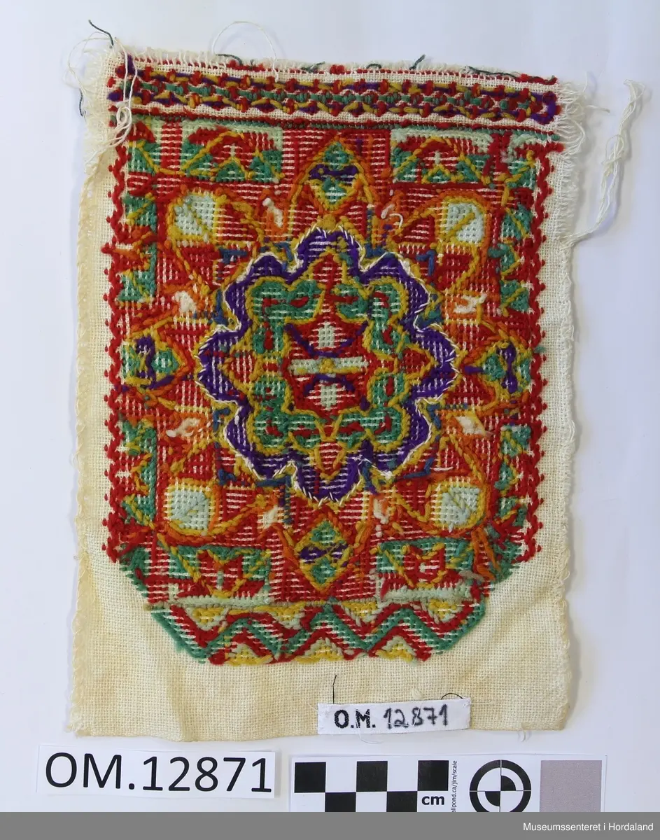 Rektangulær brystduk brodert på eit vevd på eit bomullstoff. Plattsaum (halve kross-sting). Blomstermotiv i midten sydd i fleire retningar. Brodert bord oppe og nede. Raud er hovudfargen.