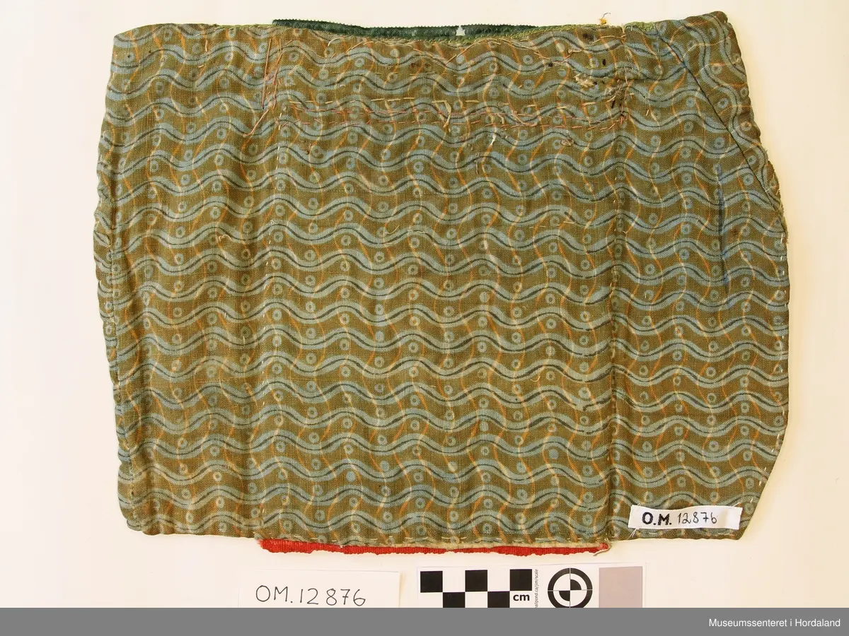 Rektangulær bringeduk med hjertemotiv i gullfarga stråperler på raudt ullstoff. Åttebladrose i stråperler inni hjertet.  Grøn kant oppe med gullfarga metallband. Sikksakk-borde med gullfarga stråperler mellom. Nokre store glasperler til dekor innimellom. Avstiva med papir og monteert på naturfarga  linstoff. Baksida av mønstra bomullstoff.