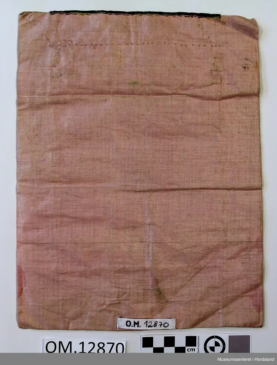 Rektangulær bringeduk med raudt ullstykke framme uten brodering. Oppe er det ein kant med gullfrga metallbord oppå eit brokadeband, metallborden går og eit stykke ned på det raude stoffet. Rosa, blankt fõrstoff til brei kanting og bakstykke. Alt er handsauma. Eit stykke papir el l inni til forsterking. Litt felkkete på forstoffet.