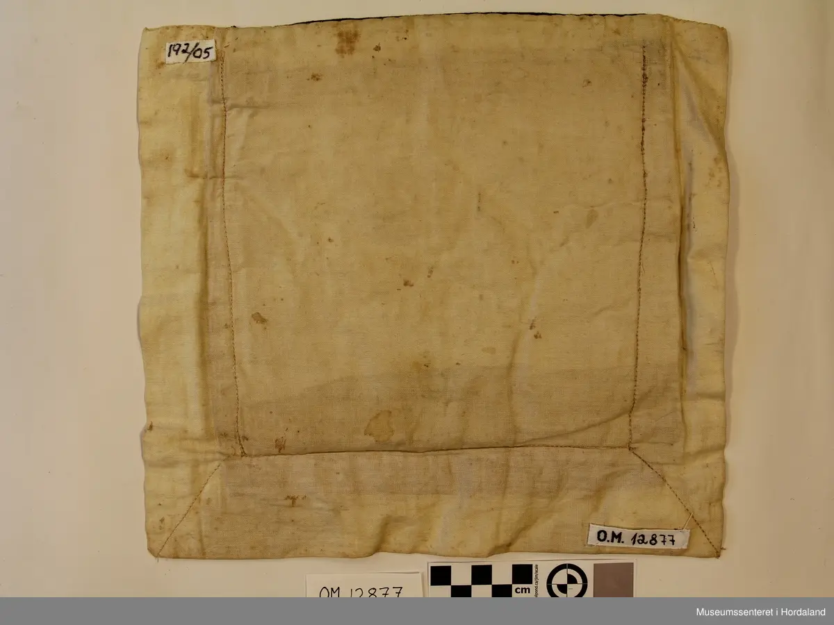 Kvadratisk bringeduk med ei heil og to halve åttebladroser i perler på raudt ullstoff. Mørkegrønt fløyelskant oppe med sikk-sakk bord stråperler. Bomullstoff til bakstykke. Montert med maskinsaum. Innlagt tynn papp som forsterking.