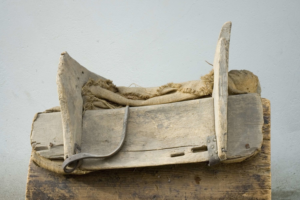 Klövjsadel med sido- och tvärstycken av trä. Mittdelen består av säckväv, stoppad med krollsplint. Båda tvärstyckena (eller valven) har en toppig form, det bakre är avhugget och det främre har ett v-format urtag. Nertill på tvärstyckena finns avlånga hål, troligen för remmar. Fyra korta spännremmar finns fortfarande kvar på sadeln. I bakre tvärstycket finns två hål där det troligen suttit en bygel.