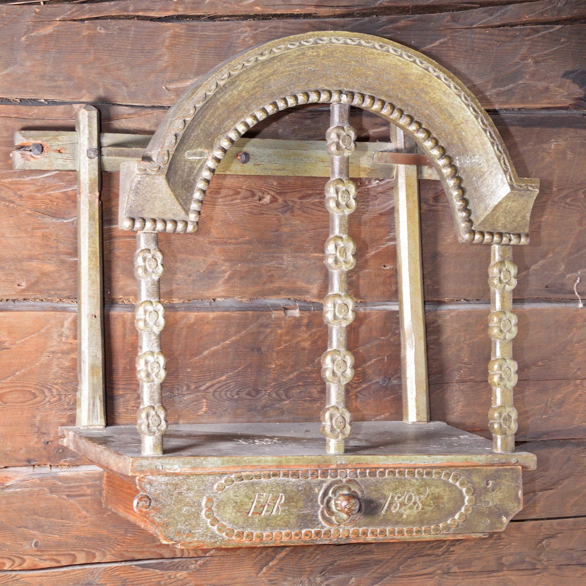 Tavlett av grönmålad furu med låda. Bågformigt krön med skuren pärlstav, uppburet av tre pelare med utskurna blommor. Låda med blomma och svarvad dragknopp. Lådfrontens hörn i form av enkel volutformation, högra hörnet saknas dock.