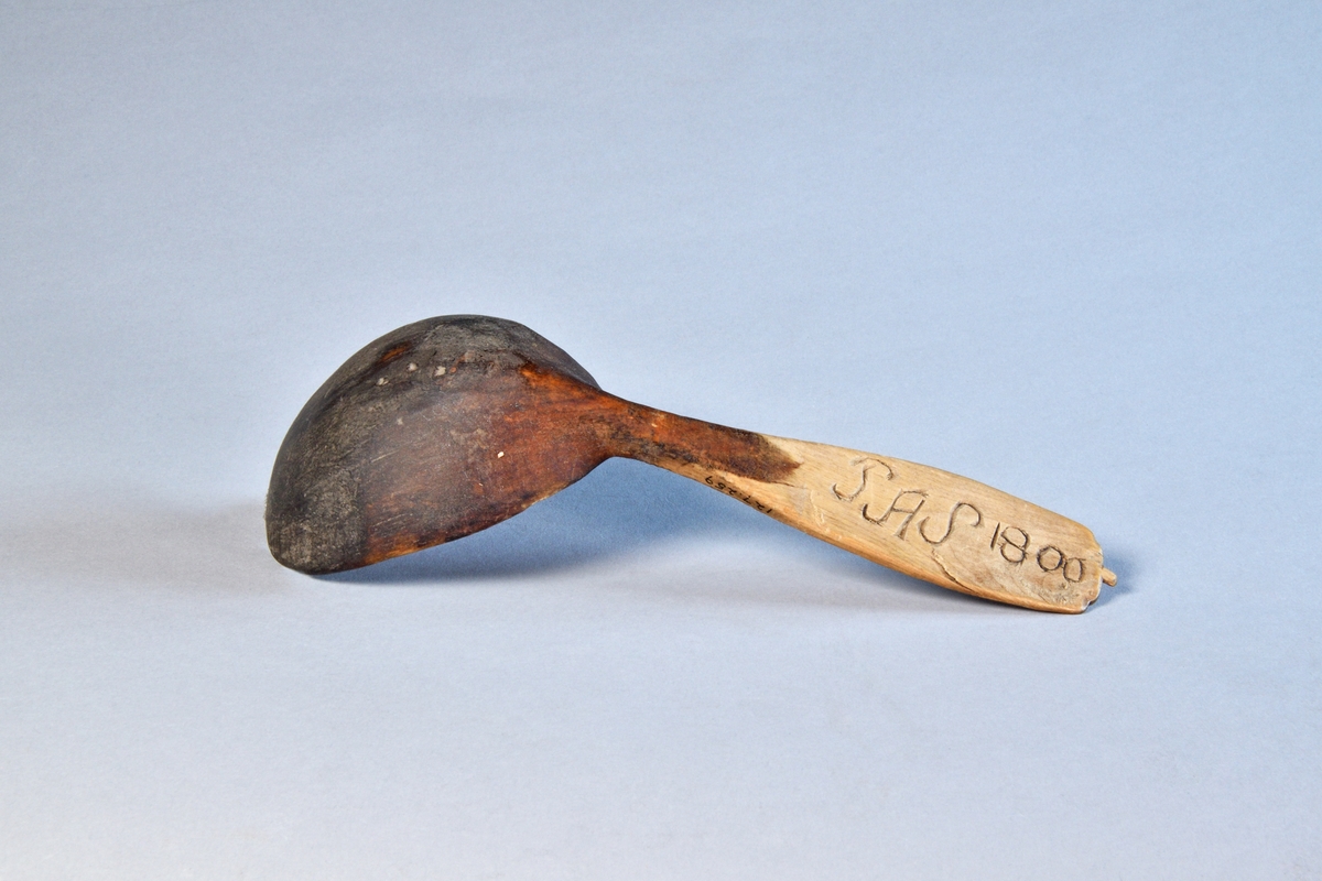 Sked av lövträ med riklig karvsnittsornamentik på skaftet. Djupt blad, böjt mot skaftet. Spår av grön bemålning på bladet.