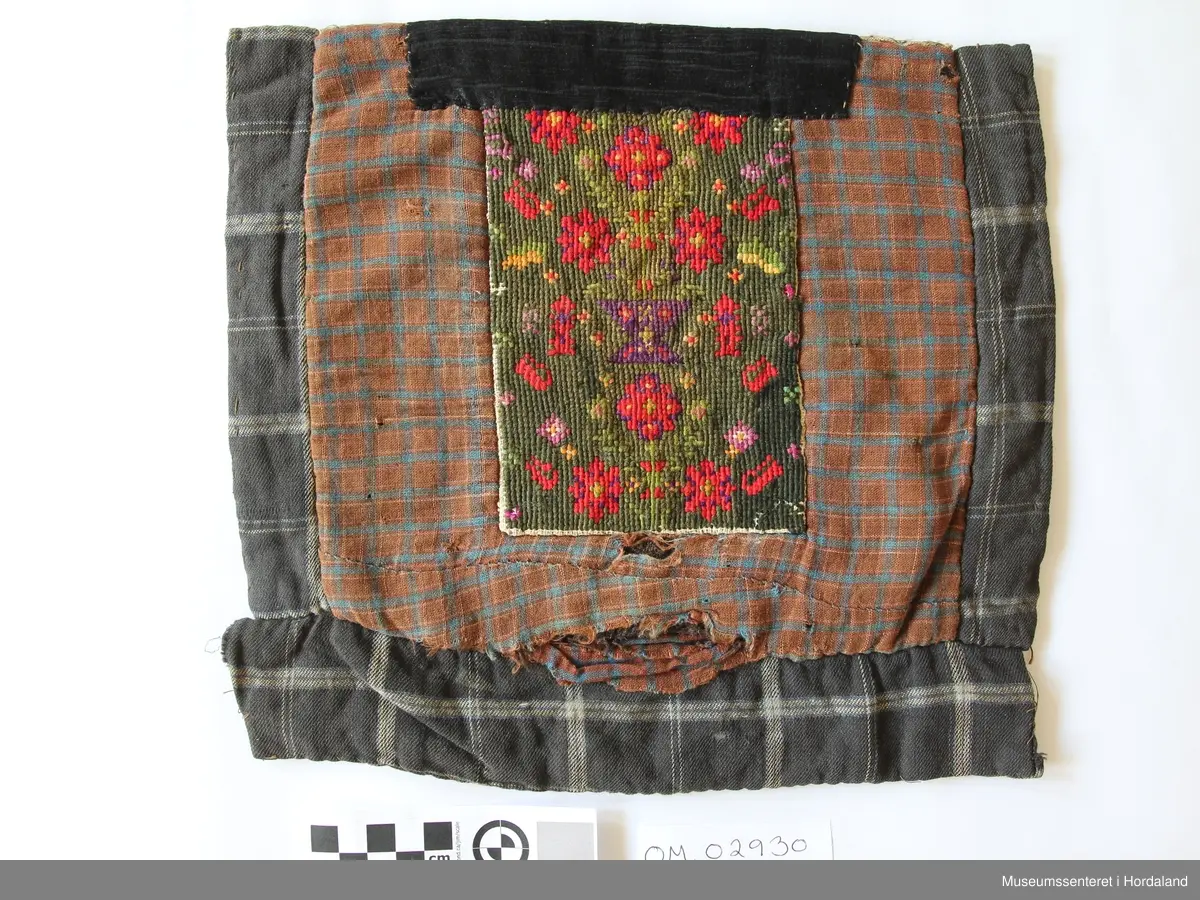 Rektangulær brystduk på kvadratisk botnstykke. Brystduken er brodert med halve kross-sting på stramei. Blomstermotiv i mange skarpe fargar. Ein svart silkeband oppe. Bomullstoff til botnstoff, bakstykke og sidestykke og stykke nederst. To forskjellige stoff til botn stoff med ruter. Brystduken er forsterka inni med tjukk kypertstoff.