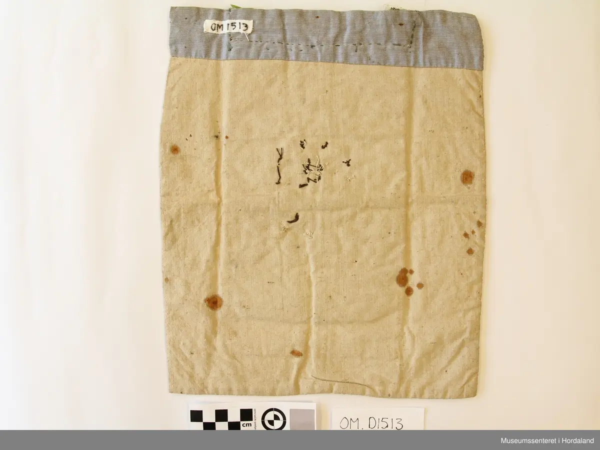 Bringeklut brodert på stramei med kross-sting og dragesaum. Raud med blått, gult, grønt og gull. Festa på eit kvitt stoff. Stoffet har ein stor flekk og nokre små. Oppe på baksida er montert på eit stripete stoff som går eit lite stykke ned på framsida. Grønt silkeband oppe med gullfarga metallkronekvarde sydd utanpå. Store og små perler sydd på broderimønsteret, nokre av desse er knuste.