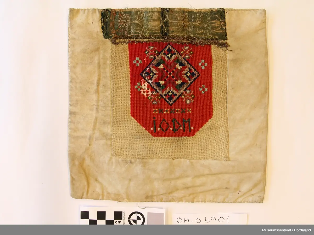 Rektangulær bringeduk sydd på kvadratisk bomullstoff. Broderiet er i halve og heile kross-sting sydd på linstoff. Raud hovudfarge. Initialane I O D M er sydd nederst på broderiet. Silkeband øvst med ei smal metallbord nedom. Silkebandet, metallborda og broderioet er veldig slitt.