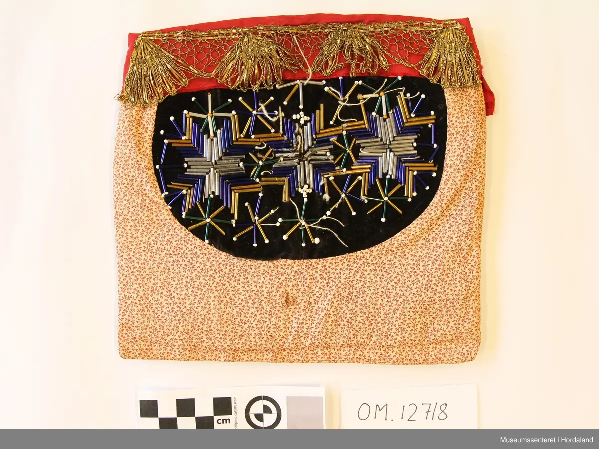 Kvadratisk bringeduk med perlemotiv med stråperler montert på svart fløyel - 3 åttebladroser. Overfor perlemotivet er det raud silkekant med gullband utanpå. Alt er montert på eit småmønstra bomullstoff som ligg som ei "lomme"  - inni "lomma" ligg eit tjukt mellomfõr.