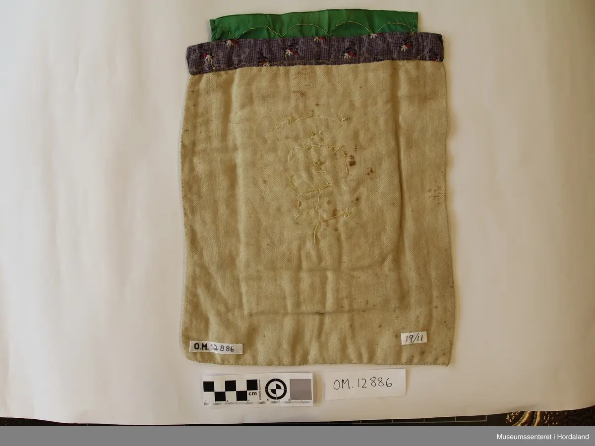 Kvadratisk bringeduk med motiv i kross-sting - hovudmotivet er dekorert med nokre store perler (to knuste). Metallkvarde oppe på eit grønt silkeband. Lilla band bakom oppe. bakstykke på eit stoff som er naturfarga.