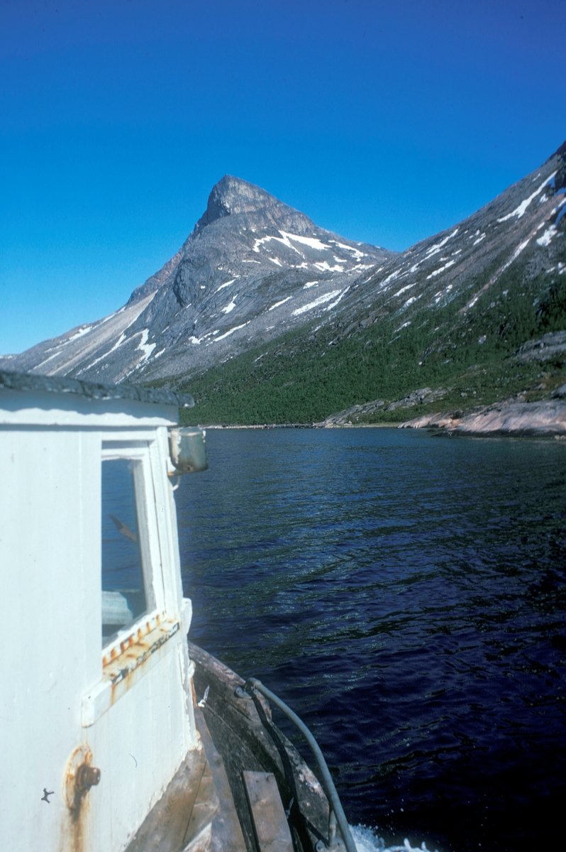 Flakstadvåg, 1976 : I forgrunnen, deler av styrhuset på en båt. I bakgrunnen ligger det et knapt synlig oppdrettsanlegg ved foten av høye fjell.