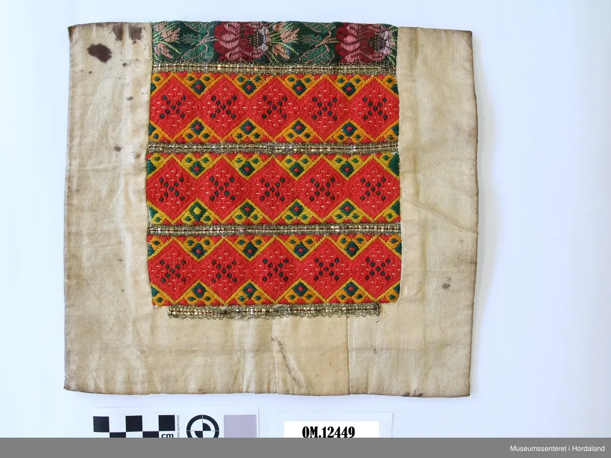 Bringeklut med ullbroderi i smøyg i fargane raudt, grønt og gult. Smale metallborder innimellom broderiet. Rosete silkeband oppe i grønt, gult og to raud/lilla fargar. montert på kvitt bomullsstoff som no er veldig gulna og flekkete. Bakstykket er i grønt vadmål.