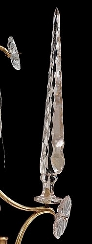 Ljuskrona med stomme uppbyggd av volutformade brännförgyllda mässningsstavar. Upptill en plymliknande konstruktion dekorerad med lövformade slipade prismor och gjutna margueriter. Ljuskronans stomme har sex ljusarmar fästa i en järnplatta med täckbricka av förgylld mässingsplåt. Ljusarmen avslutas med gjutna ljuspipor och droppskålar. Mittstång av järn med blåst stamglas format som ett slipat klot upptilll krönt med en obelisk. Ljusarmarna är dekorerade med lövformade slipade prismor och gjutna margueriter. Till varje ljusarm finns sex obelisker med slipade ytterkanter. Stommen är dekorerad med stavverk av glasrör och glaspärlor trädda på metalltråd. Nedtill hänger ett större glasklot med slipad dekor.