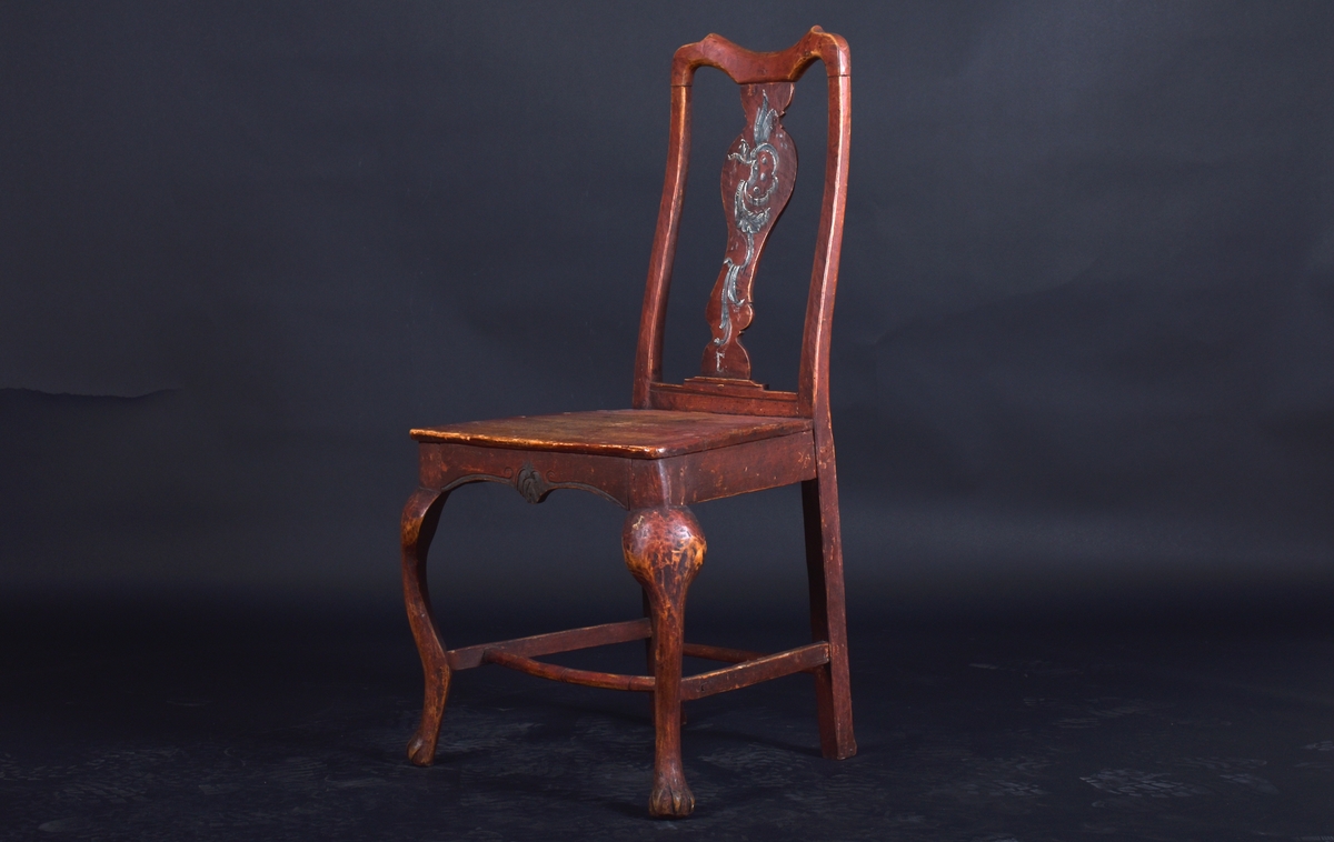 Stol av björk och furu, på fyra svängda ben. Ryggen har en mittbricka i balusterform. Stolen är målad i mörkt rött och ryggbrickan har målad dekoration i rokokostil i grått och vitt.